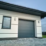 Nowoczesny garaż z szarą bramą segmentową, białymi ścianami i kamienną podłogą.
