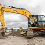 Żółta koparka JCB na placu budowy, pracownik w kamizelce odblaskowej przy gąsienicy.