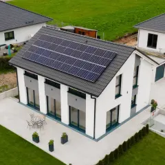 Nowoczesny dom z panelami słonecznymi na dachu, otoczony zielenią i podjazdem.