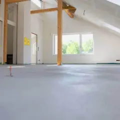 Nowe wnętrze z drewnianymi belkami i oknem. Na podłodze świeża wylewka, która czeka na wykończenie. Zastanawiasz się, ile cementu na 100m2 wylewki?
