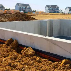 Betonowe ściany fundamentowe schną, czekając na dalsze prace budowlane. W tle widać nowe domy.