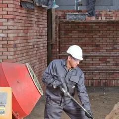 Budowlaniec w kasku pracuje przy betoniarce. Obok stos piasku.