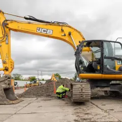 Żółta koparka JCB na placu budowy, pracownik w kamizelce odblaskowej przy gąsienicy.