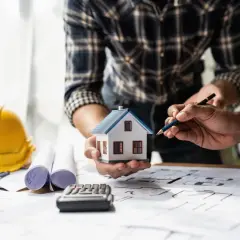 Architekci omawiają projekt domu, planując, jak uzyskać pozwolenie na budowę. Na stole makieta domu, plany, kask ochronny i kalkulator.