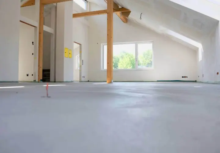 Nowe wnętrze z drewnianymi belkami i oknem. Na podłodze świeża wylewka, która czeka na wykończenie. Zastanawiasz się, ile cementu na 100m2 wylewki?