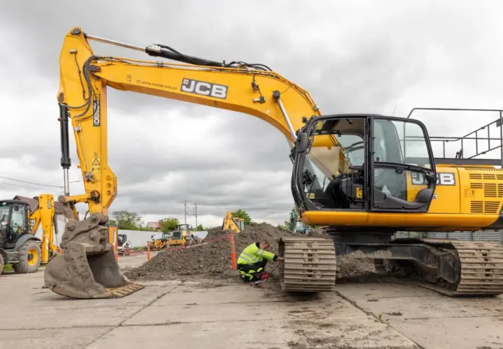 Żółta koparka JCB na placu budowy, pracownik w kamizelce odblaskowej przy gąsienicy.