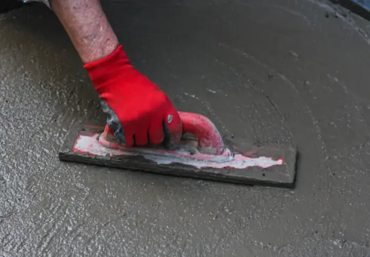 Ręka w czerwnej rękawicy wygładza mokrą wylewkę samopoziomującą do garażu za pomocą kielni.