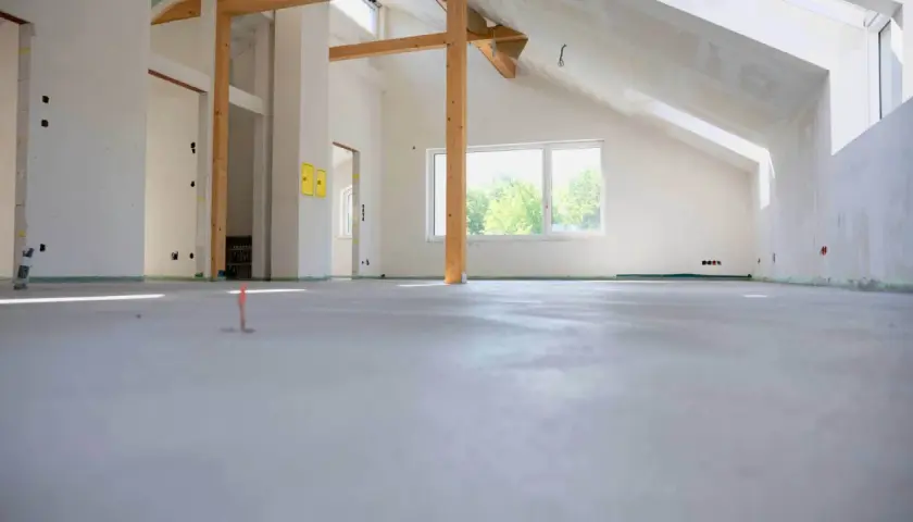 Nowe wnętrze z drewnianymi belkami i oknem. Na podłodze świeża wylewka, która czeka na wykończenie. Zastanawiasz się, ile cementu na 100m2 wylewki?