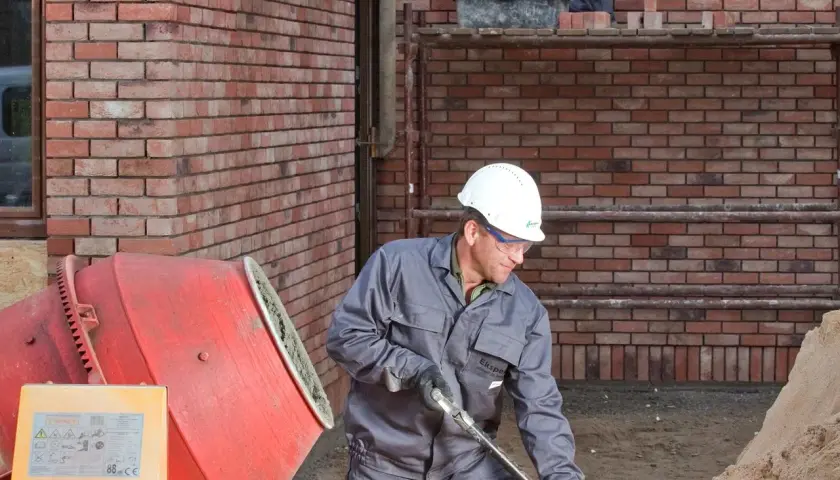 Budowlaniec w kasku pracuje przy betoniarce. Obok stos piasku.