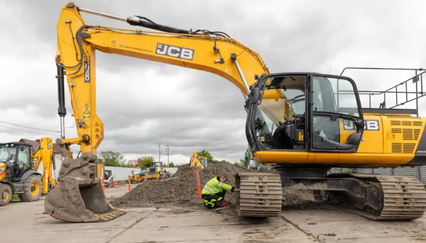 Żółta koparka JCB na placu budowy, pracownik w kamizelce odblaskowej przy gąsienicy.