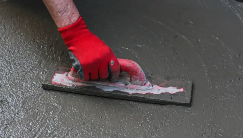 Ręka w czerwnej rękawicy wygładza mokrą wylewkę samopoziomującą do garażu za pomocą kielni.