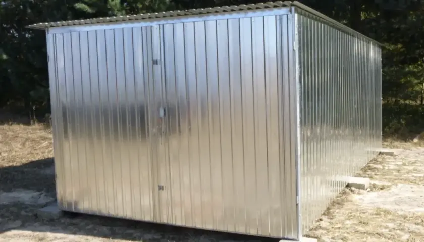 Nowy garaż blaszany z dwuskrzydłowymi drzwiami, stojący na betonowych bloczkach, na tle drzew.
