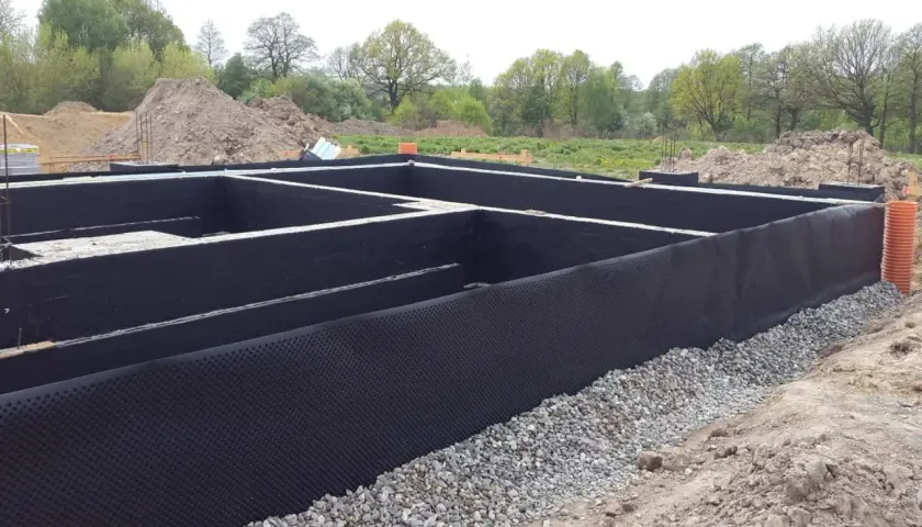 Fundament domu w budowie, z czarną izolacją i otoczakami.