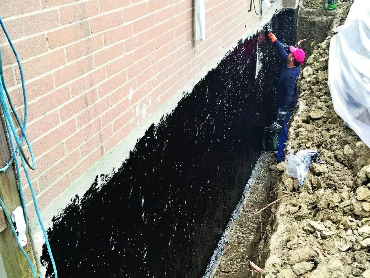 Pracownik nakłada czarną hydroizolację na ścianę fundamentową.