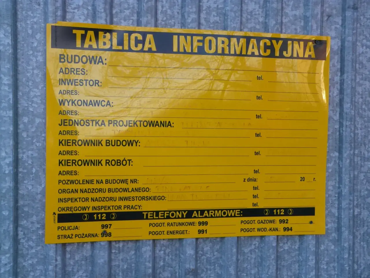 Tablica informacyjna na budowie. Widać pola na dane inwestora, kierownika budowy, numer pozwolenia na budowę i telefony alarmowe. Ważne jest pozwolenie na budowę, by wszystko było legalne.