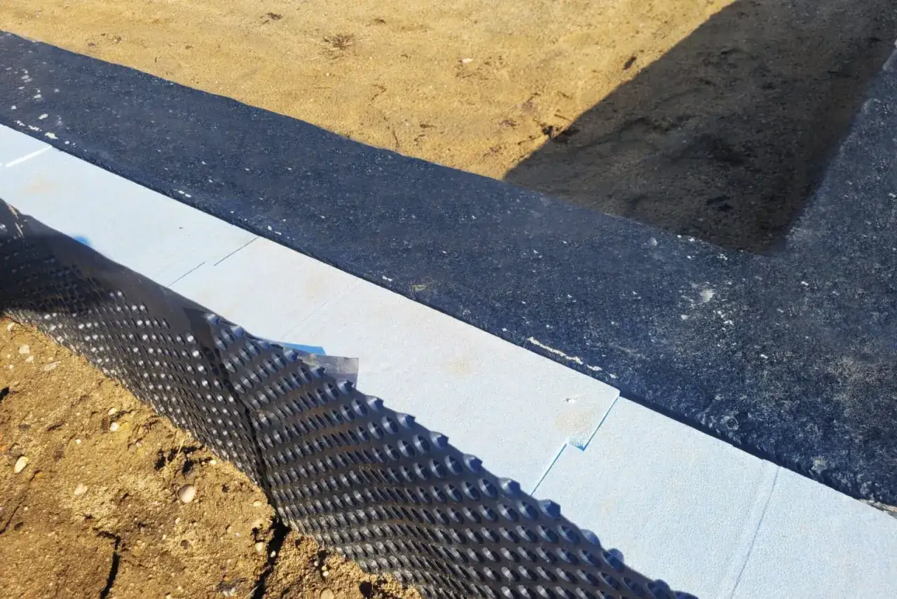 Płyty z jasnoniebieskiego styropianu, idealne jako jaki styropian na fundamenty, są układane obok czarnej membrany izolacyjnej.