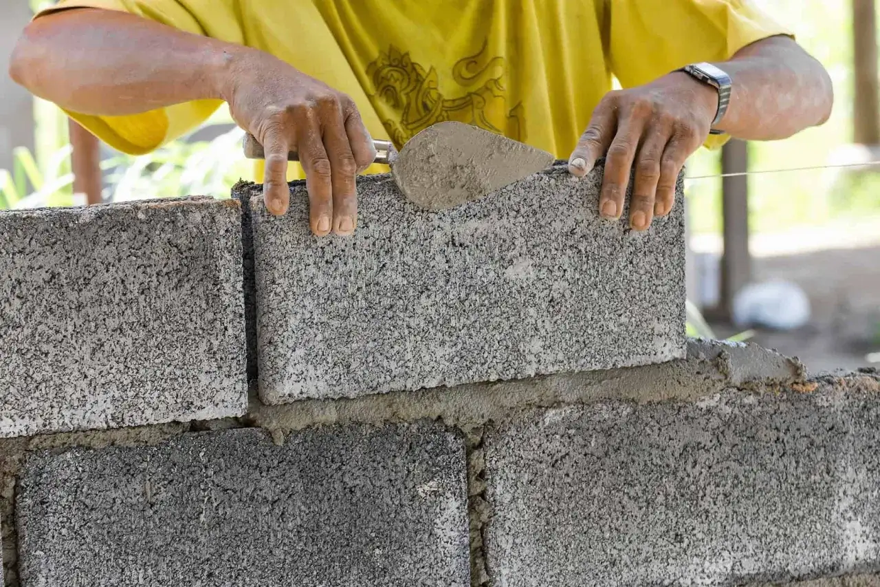 Budowniczy układa bloczki na fundament, używając kielni do zaprawy. Widać, ile bloczków na fundament potrzeba do budowy.